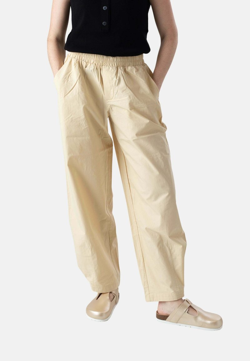 Persona con pantalones beige de pierna ancha con cintura elástica, camiseta negra sin mangas y zuecos beige de poner y quitar, de pie con las manos en los bolsillos.