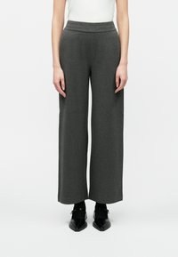 Žena v šedých širokých nohaviciach vysokého pásu, s čiernymi špicatými Mary Jane topánkami a bielym tričkom, stojí na pozadí bielej steny.