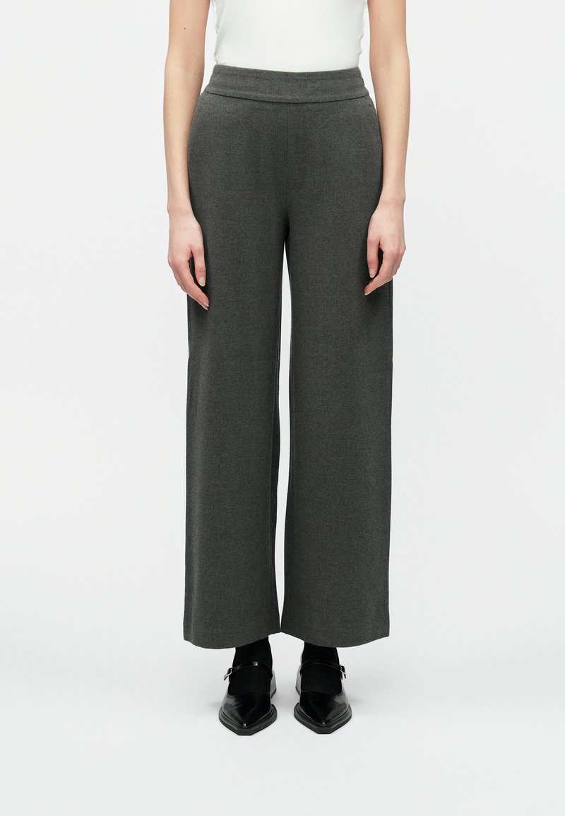 Žena v šedých širokých nohaviciach vysokého pásu, s čiernymi špicatými Mary Jane topánkami a bielym tričkom, stojí na pozadí bielej steny.