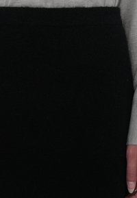 Personne portant une chemise grise à manches longues et une jupe noire, main visible sur le côté, debout devant un fond uni.