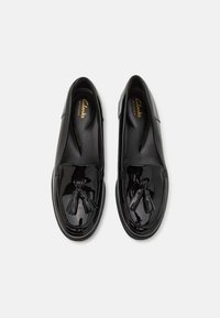 Black patent leather slip-on shoes with a rounded toe and decorative tassels on the vamp. Flat sole and cushioned insole for comfort.