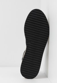 Sole černé boty s vodorovnými drážkami, velikost 37, umístěná svisle proti rozhraní bílé zdi a podlahy.