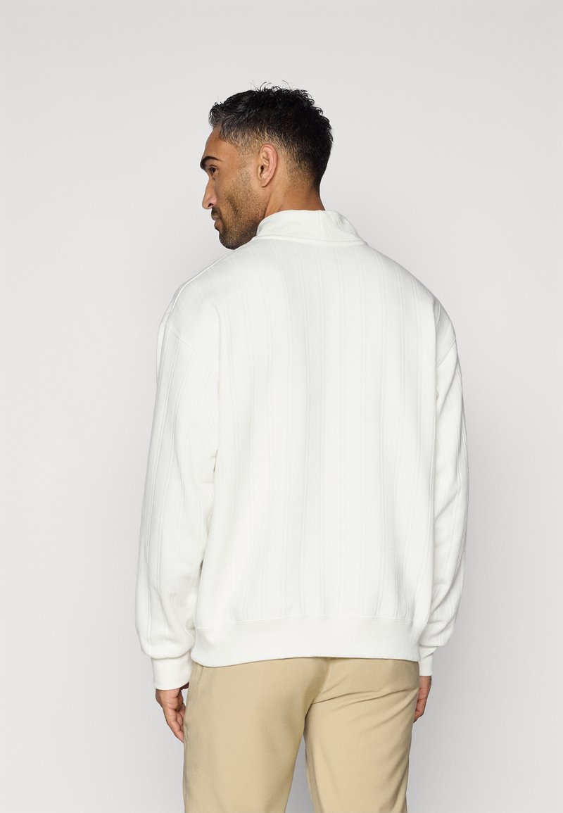 Weißer Pullover mit gerippter Struktur, hohem Kragen und lockerer Passform. Hat lange Ärmel und einen Saum, der auf der Taille sitzt.