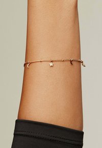 Pulsera de cadena de oro con encantos de luna creciente y estrella, con pequeñas cuentas redondas, exhibida en un brazo bronceado que lleva una manga negra.