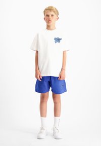 Witte katoenen t-shirt met blauwe grafische print; gecombineerd met blauwe shorts met een logo. Witte sneakers en witte sokken maken de outfit compleet.