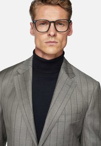 Veste de costume gris à rayures avec revers en pointe, portée avec un col roulé noir. Le mannequin porte des lunettes noires, mettant en avant un look moderne et cintré.