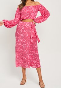 Růžový crop top bez ramínek s balónovými rukávy a odpovídající sukni se zavázáním v pase. Oba mají bodkovaný vzor a jsou z lehkého materiálu.