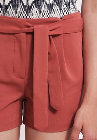 Pantaloni corti di colore ruggine realizzati in tessuto liscio, dotati di una vita con lacci e tasche laterali. Il design è sartoriale, con linee pulite.