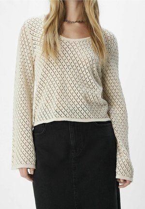 Frau trägt einen cremefarbenen, langärmligen Strickpullover mit Rautenmuster und einen schwarzen, hoch taillierten Rock, steht vor einem schlichten Hintergrund.