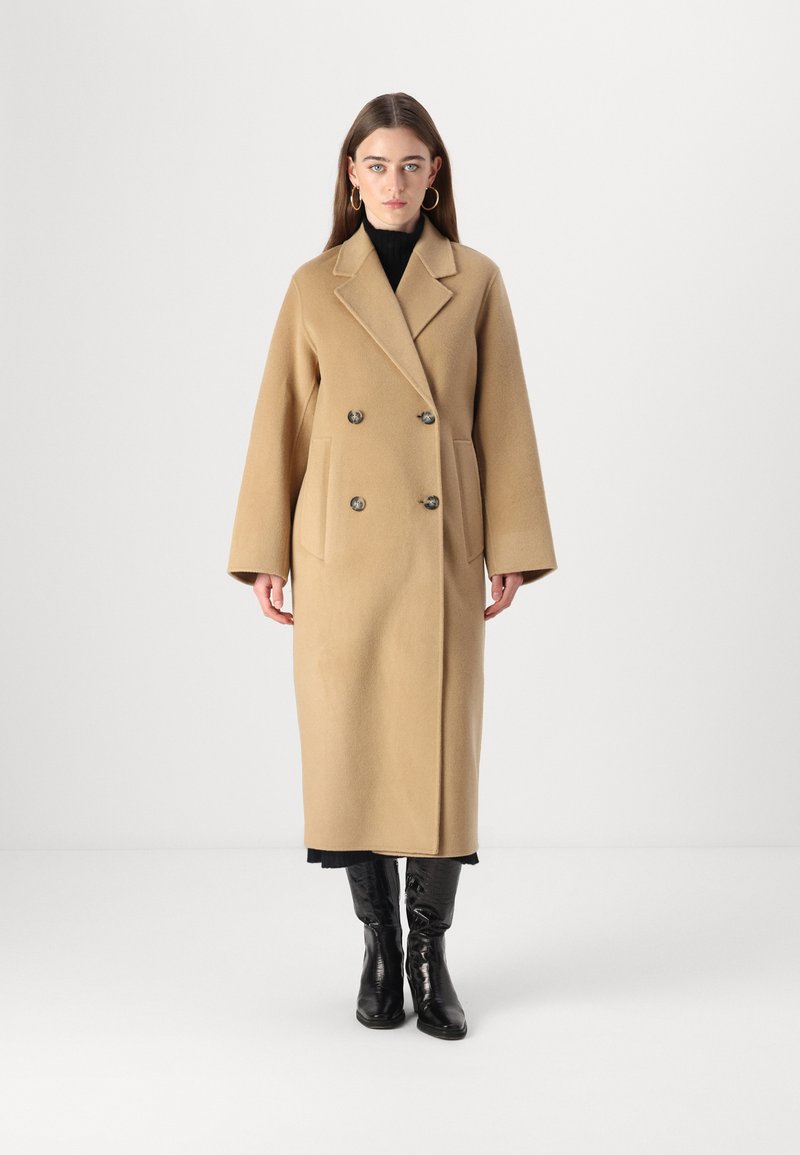 Cappotto beige in misto lana, doppiopetto, con colletto a rever, grandi bottoni, tasche laterali e maniche ampie, che arriva a metà polpaccio.