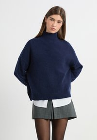 Granatowy, oversized sweter z golfem wykonany z miękkiego materiału, z prążkowanymi mankietami i dołem, noszony na białej koszuli z kołnierzykiem i szarej spódnicy.