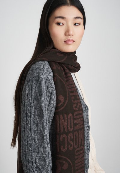 Jonge vrouw met lang, donker haar draagt een twee-kleurige kabelgebreide cardigan en een donkerbruine sjaal met opvallende merktekst.
