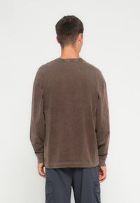 Hnedá košeľa s dlhým rukávom a okrúhlym výstrihom, jemná textúra a voľný strih. Obsahuje viditeľné švy a detaily s nedokončeným lemovaním.