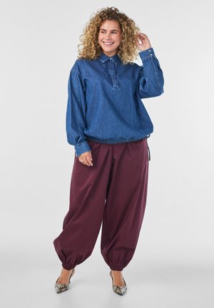 Femme souriante aux cheveux bouclés portant une chemise en jean bleue à manches longues, un pantalon ample bordeaux avec des poignets élastiques et des chaussures à talons hauts à motifs.