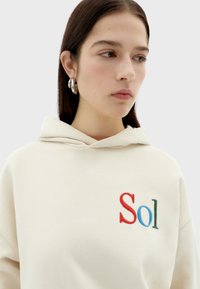 Cream hoodie with a drawstring, featuring "Sol" embroidered in red, blue, and green. Smooth texture, minimalistic design.