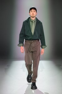 Il modello indossa una giacca verde scuro, una camicia grigio-verde e pantaloni marroni con cordino. Stivali neri completano l'outfit, caratterizzato da texture morbide e design aderente.