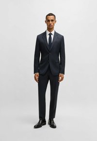 Completo blu a righe con vestibilità su misura, giacca monopetto e pantaloni coordinati. Camicia bianca e cravatta scura, abbinati a scarpe nere.
