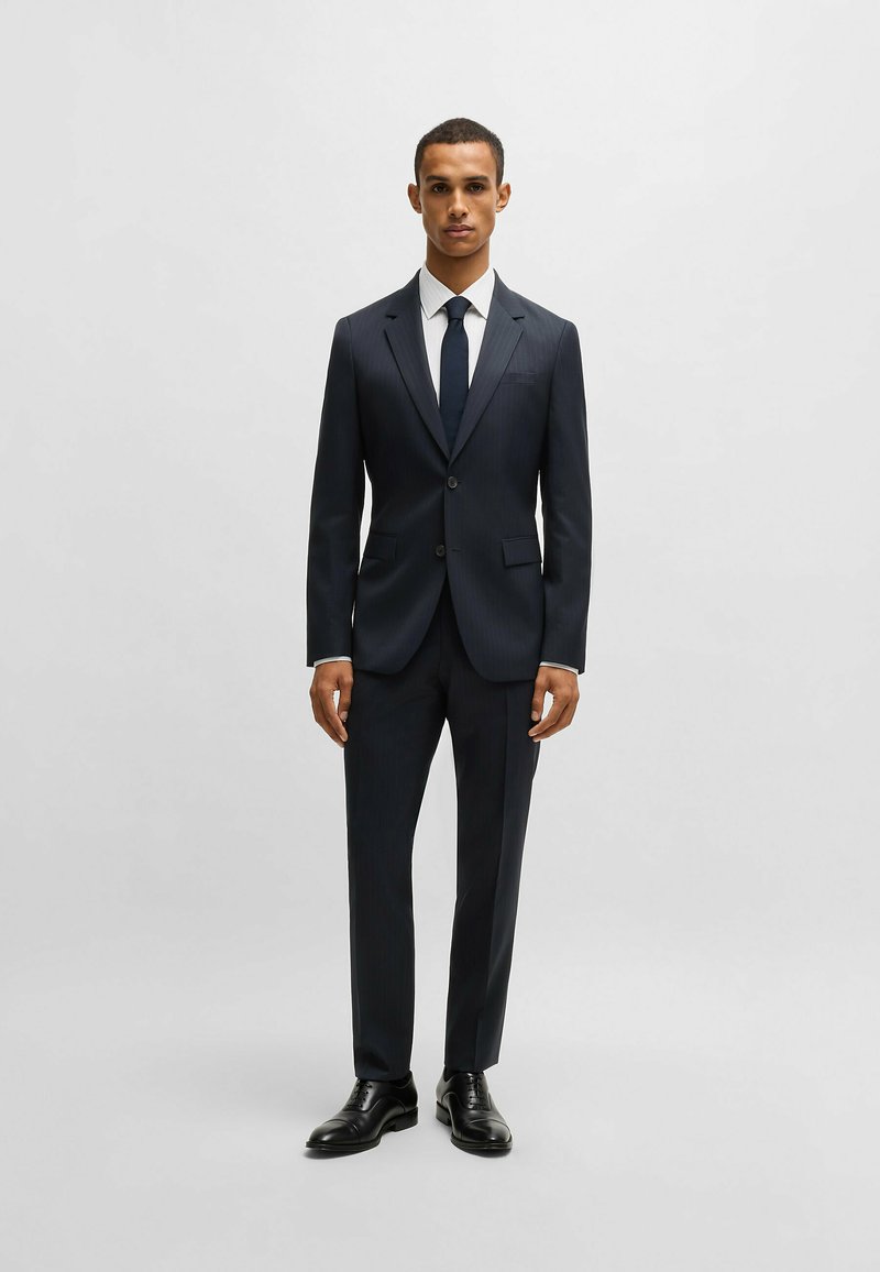 Completo blu a righe con vestibilità su misura, giacca monopetto e pantaloni coordinati. Camicia bianca e cravatta scura, abbinati a scarpe nere.
