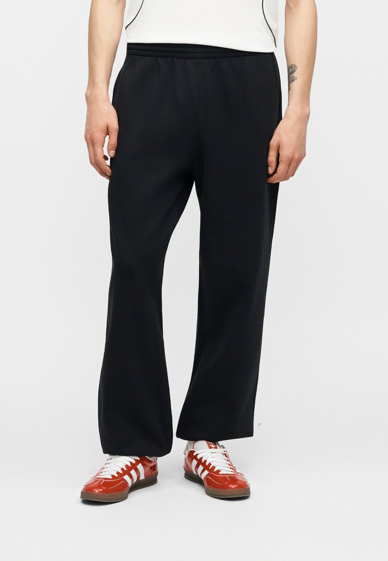 Person trägt schwarze, lockere Hose und rote Sneakers mit weißen Streifen, steht vor einem schlichten hellen Hintergrund.