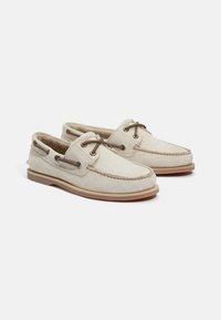 Beige Bootsschuhe mit einer weichen Wildleder-Oberfläche, Leder-Schnürsenkeln und kontrastierenden Nähten, ausgestattet mit einer Gummisohle und seitlichen Ösen zur Belüftung.