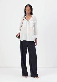 Camicia bianca trasparente con bottoni e maniche lunghe, due tasche frontali, abbinata a pantaloni neri a gamba larga e sandali minimal. Accessorio: piccola borsa.
