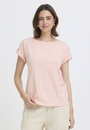 Jeune femme aux cheveux bruns mi-longs portant des boucles d'oreilles en or, un haut rose clair à manches courtes et un pantalon beige sur fond blanc.
