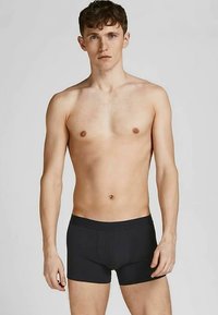 Mlad muškarac s kratkom smeškom kosom, obučen u crne bokserice, stoji nasuprot jednostavne svijetlosive pozadine, okrenut prema naprijed.