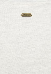 Bílá žebrovaná látka s texturovaným povrchem. Obsahuje malou bronzovou značku s vyrytým názvem "Barbour", umístěnou blízko výstřihu.