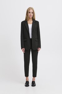 Fekete, testhez simuló blézer, bemetszett hajtókával, egysoros gombolással, fekete, szűk szabású nadrággal párosítva. A modell fehér felsőt és fényes fekete cipőt visel.