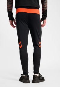 Czarne sportowe spodnie z pomarańczowym elastycznym paskiem w talii, ozdobione trzema pomarańczowymi szewronami na dolnych partiach nóg. Gładka tekstura, dopasowany krój.