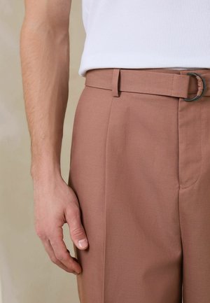 Mann trägt hellbraune Hose mit passendem Stoffgürtel und weißes, geripptes ärmelloses Shirt, Hand entspannt an der Seite.