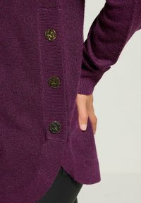 Lila Strickpullover mit einem seitlichen Knopfdetail und vier Metallknöpfen. Der Stoff hat eine weiche Textur und einen lockeren Schnitt.