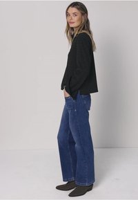 Donna in piedi di profilo con le mani nelle tasche, indossa una maglia nera a maniche lunghe, jeans blu a gamba larga e stivaletti neri su uno sfondo semplice.