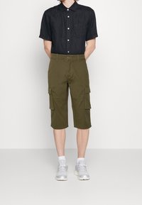 Chemise noire en lin à manches courtes, shorts cargo olive avec poches et chaussures de sport grises avec accents blancs. Design simple et coupe ajustée.