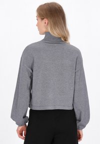 Frau mit schulterlangem blonden Haar trägt einen grauen Rollkragenpullover mit langen Ärmeln und schwarze Hosen, von hinten auf weißem Hintergrund betrachtet.