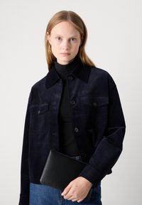 Veste en velours côtelé bleu marine avec de grandes poches avant, fermeture à boutons et col rabattu. Le mannequin tient une pochette en cuir noir.