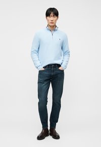 Άνδρας που φοράει ανοιχτό μπλε πουλόβερ με φερμουάρ στο λαιμό, καρό πουκάμισο, σκούρο μπλε τζιν και σκούρα καφέ δερμάτινα μποτάκια, στέκεται με τα χέρια στις τσέπες.