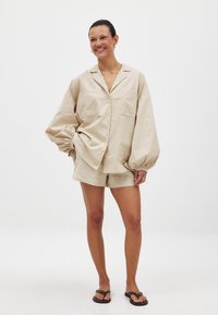Beige linne skjorta med överdimensionerade, puffiga ärmar och två framfickor, i kombination med matchande shorts och svarta flip-flops.