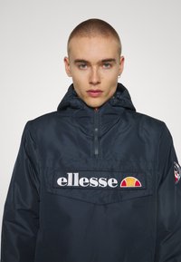 Giacca con cappuccio blu navy con zip a un quarto, caratterizzata da una grande toppa logo sul davanti e un emblem sulla manica sinistra. Tessuto liscio e leggero.