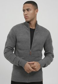 Grauer gestrickter Cardigan mit einem Reißverschluss, gerippten Bündchen und Saum, strukturiertem Muster und Stehkragen. Auffällige Kontrastnähte.