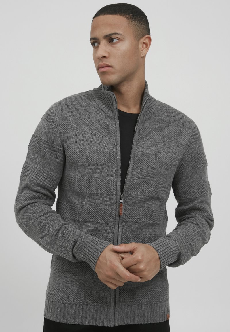 Grauer gestrickter Cardigan mit einem Reißverschluss, gerippten Bündchen und Saum, strukturiertem Muster und Stehkragen. Auffällige Kontrastnähte.