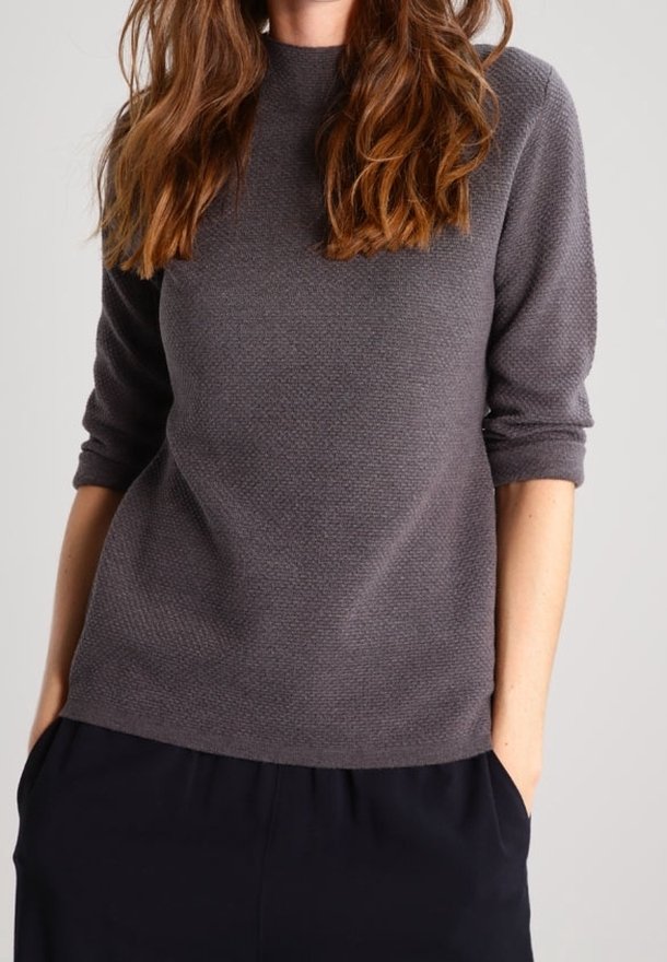 Femme aux longs cheveux bruns portant un haut gris texturé à manches longues et un pantalon noir, les mains dans les poches, sur un fond uni.