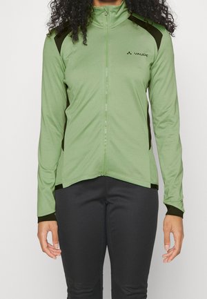 Frau trägt eine hellgrüne Jacke mit Reißverschluss, schwarzen Seiten- und Schulterpaneelen sowie schwarze Hose, steht vor einem einfarbigen Hintergrund.