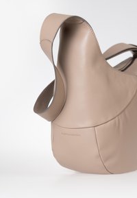 Beige Tasche aus weichem Leder mit breitem Tragegurt, abgerundetem Boden und dezentem geprägtem Markenlogo auf glatter Oberfläche.