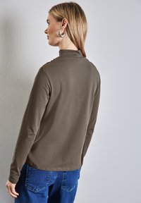 Pitkähihainen ruskea paita, jossa on korkea kaula-aukko, pehmeä rakenne ja hieman rento istuvuus, yhdistettynä sinisiin farkkuihin. Ommeltu sauma helmassa näkyvissä.