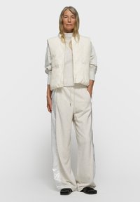 Hellfarbige gesteppte Weste über einem grauen Sweatshirt, kombiniert mit lockeren, hellgrauen Sweatpants mit glänzenden Seitenstreifen. Schwarze Schuhe.