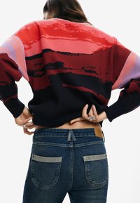 Maglione oversize rosso e blu navy con motivi astratti, abbinato a jeans in denim scuro con dettagli in grigio chiaro sulle tasche e una toppa in pelle.