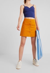 Modrá top s volánky, oranžová sukně s knoflíky vpředu, bílé tenisky. Sukně má přímý tvar a hladký materiál, zatímco top je upnutý.