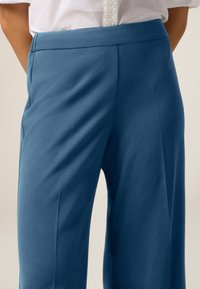 Pantaloni su misura in tessuto blu scuro, con una texture liscia, vestibilità slim e un fronte piatto con pieghe frontali per un tocco di dettaglio.