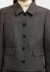 Harmaa räätälöity bleiseri, jossa on kaulus, kuusi mustaa nappia, kaksi etutaskua ja teksturoitu kangas. Yksityiskohtiin kuuluvat istuva muotoilu ja siistit linjat.